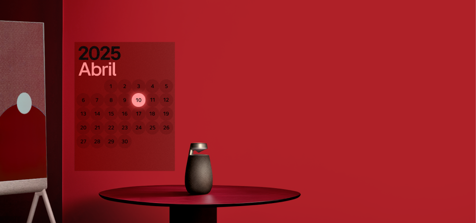 붉은색 배경의 공간에 2025년 4월 달력이 표시되어 있다. “2025 Abril”이라는 제목 아래 원형으로 배열된 날짜들이 보이고, 10일이 밝게 강조되어 있다. 오른쪽에는 둥근 검은 스피커가 테이블 위에 놓여 있으며, 왼쪽에는 스탠드형 디스플레이 일부가 보인다.